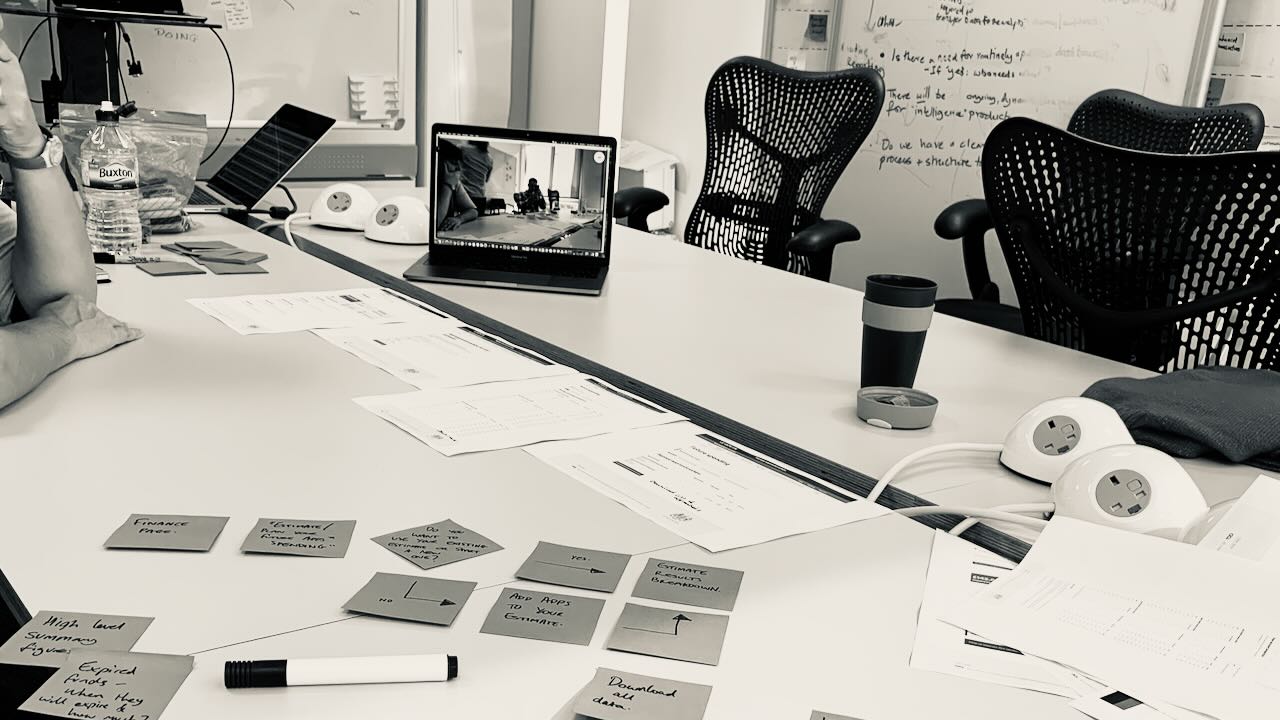 A photo of a workshop with the digital team at the Apprenticeship Service. The team are sat around a table, with a whiteboard behind them, a laptop and post-it notes are on the table.